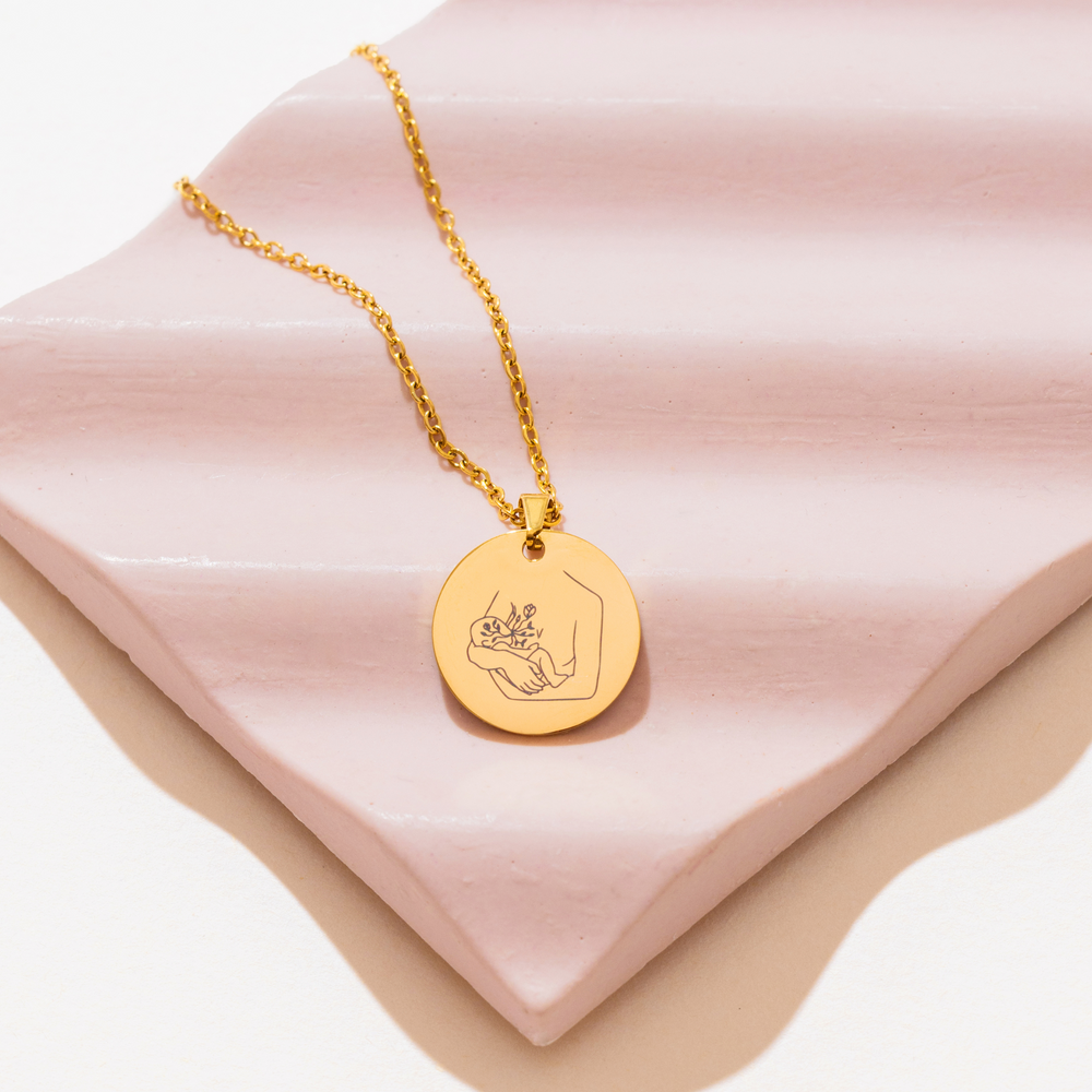 [main] A gold  pendant necklace with a gold chain featuring a mother breastfeeding her child. 