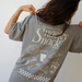[main] A mom wearing a grey T-shirt with the phrase "if you're reading this bring me a little snacky snack mmmkay" while featuring an assortment of snacks in white font on the back of the T-shirt. 