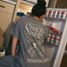 [main] A mom wearing a grey T-shirt with the phrase "if you're reading this bring me a little snacky snack mmmkay" while featuring an assortment of snacks in white font on the back of the T-shirt. 
