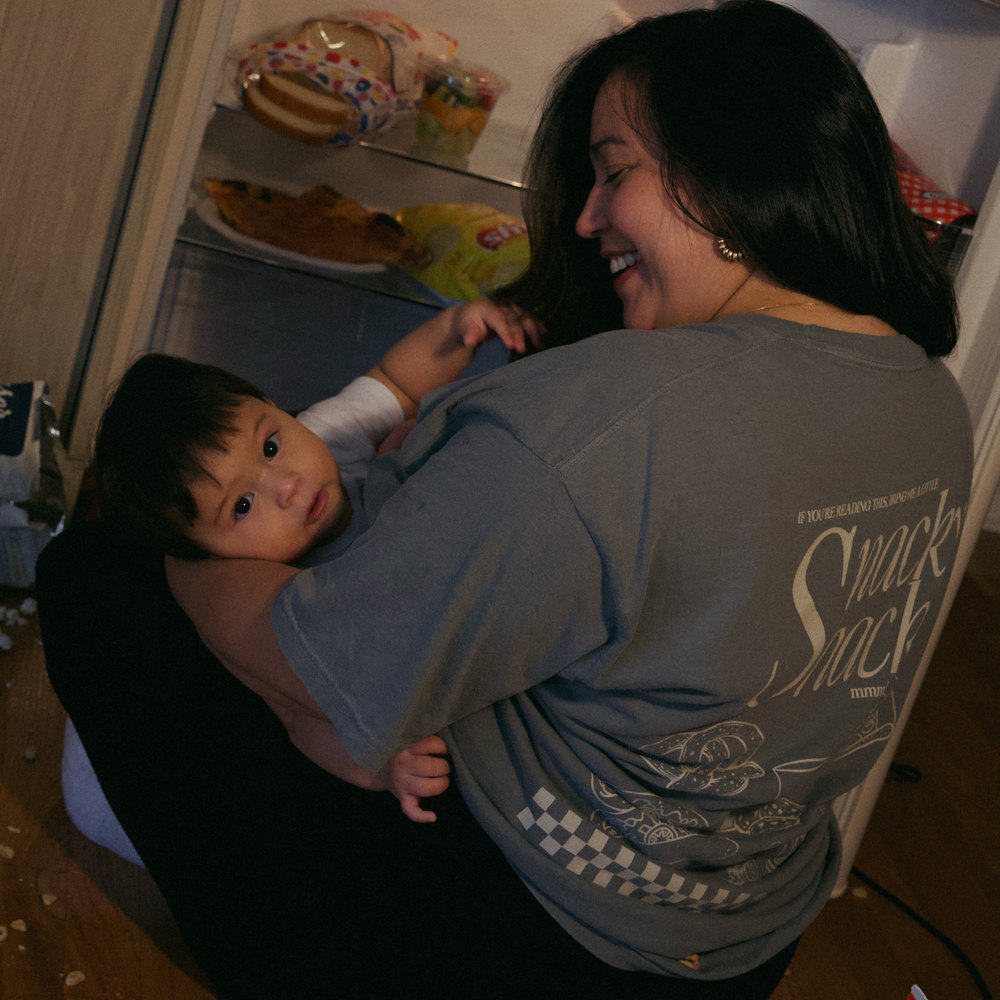 [main] A mom wearing a grey T-shirt with the phrase "if you're reading this bring me a little snacky snack mmmkay" while featuring an assortment of snacks in white font on the back of the T-shirt, with her son by the fridge.