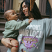 [main] A mom wearing a light sage t-shirt featuring the slogan Support Your Local Breastfeeding Mom and a bright cereal bowl graphic, while feeding her son a piece of pizza.