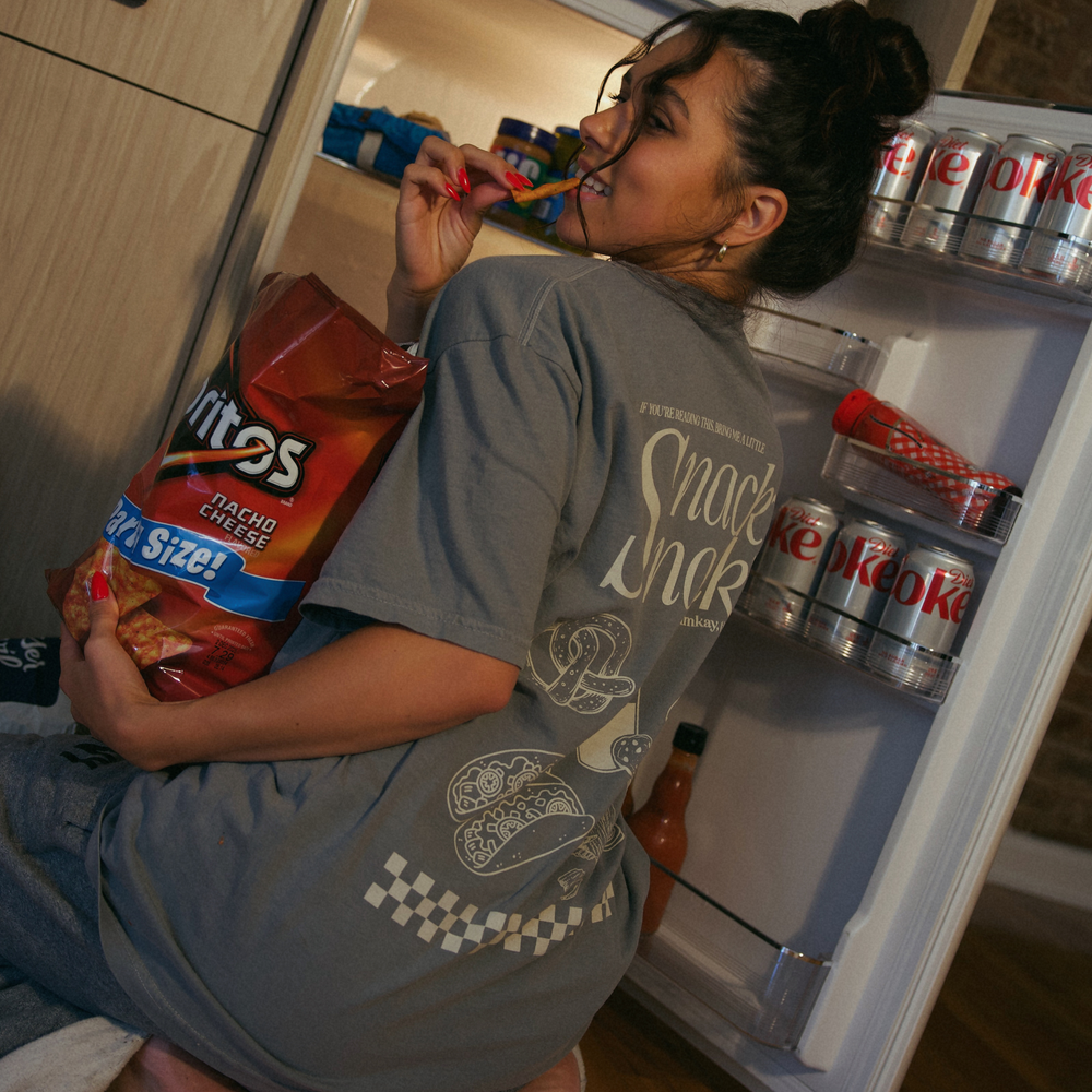 [main] A mom wearing a grey T-shirt with the phrase "if you're reading this bring me a little snacky snack mmmkay" while featuring an assortment of snacks in white font on the back of the T-shirt, while eating chips by a fridge.